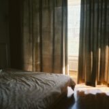 Dormitor liniștit, cu lenjerie albă și lumină blândă de dimineață pătrunzând prin draperii
